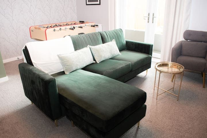 Living room with large comfortable L shaped sofa, additional chair, 65" Smart TV, football/foosball table and small table with stools.