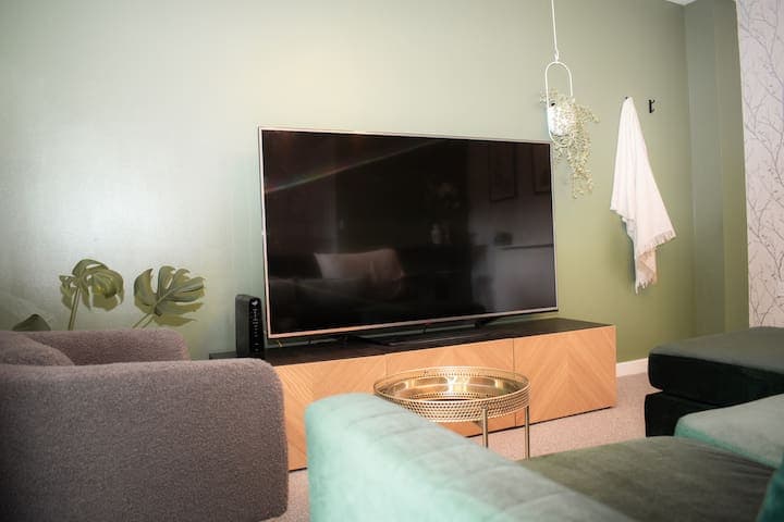 Living room with large comfortable L shaped sofa, additional chair, 65" Smart TV, football/foosball table and small table with stools.