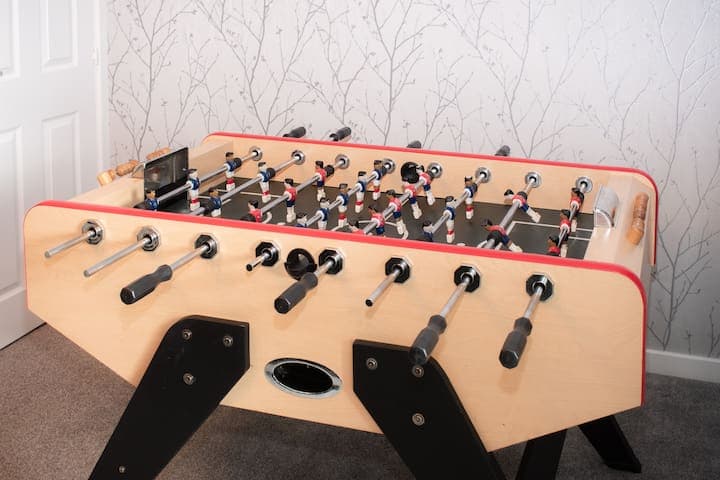 Living room with large comfortable L shaped sofa, additional chair, 65" Smart TV, football/foosball table and small table with stools.