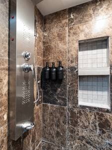 Cozy Guest Bath | Modern Shower Features the same dark marble tiles as the main bath, with a chrome-finish showerhead offering 5 settings. Includes a double built-in niche and dispensers for shampoo, conditioner, and body wash for your convenience.