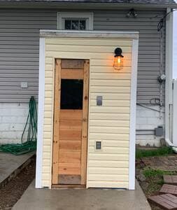 Backyard | Sauna Exterior View Sauna features home-style siding, a paneled door with a window, and an exterior light (seen upper right). An outdoor outlet is also visible.